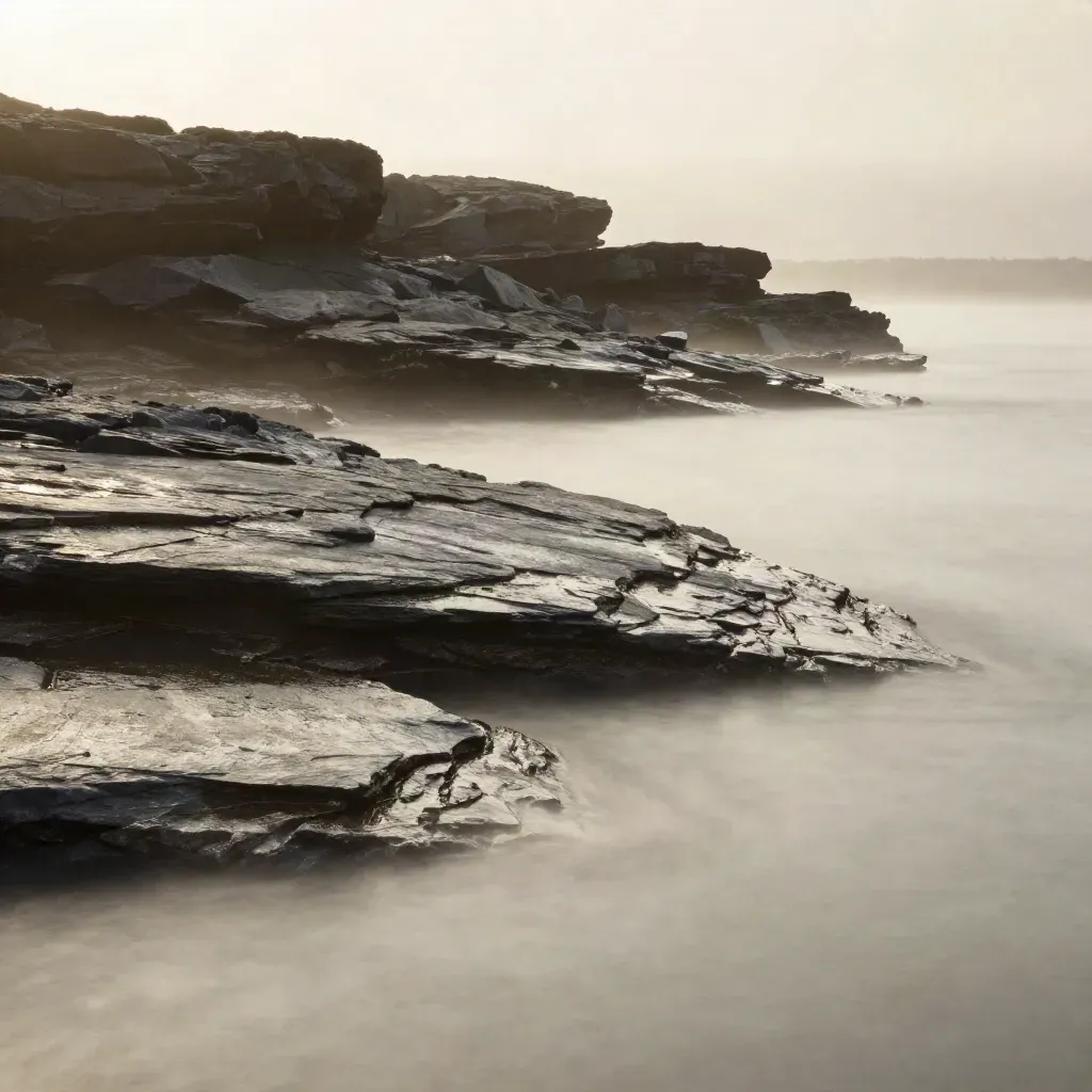 Misty coast with rock shelves, silver inlet water, and a distant slate headland at dawn
